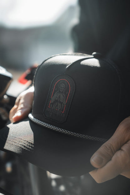 A person holds the Saints and Sinners Hat by Born United Private Label, a black cap with an embroidered cloaked figure, red outline, and decorative front chain. Soft lighting highlights this mysterious piece.