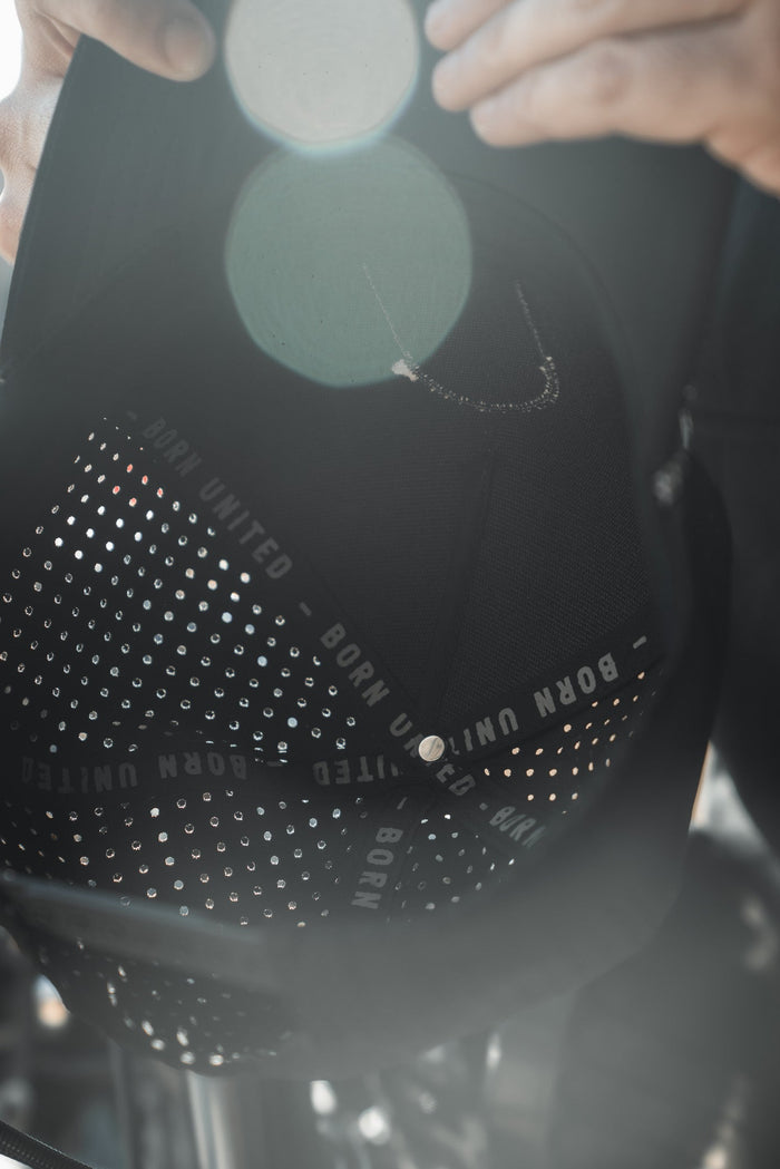 A close-up image shows the Saints and Sinners Hat from Born United Private Label, featuring BORN UNITED in white on the inner band. The black cap is dotted with small perforations, accentuated by lens flares. A partially visible person holds it reverently, cherishing the sense of unity it represents.