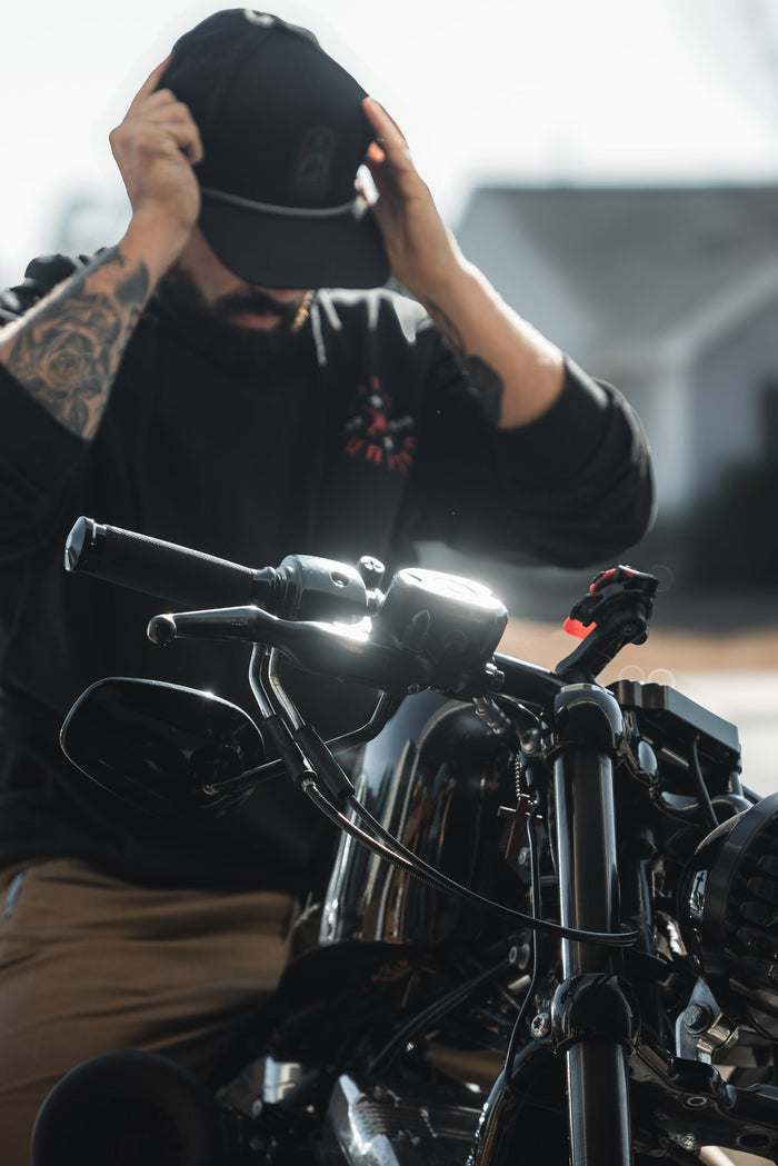 A tattooed man, embodying the spirit of a modern-day saint, adjusts his Saints and Sinners Hat by Born United Private Label while sitting on a motorcycle. The bikes handlebars are in focus as sunlight casts reflections on its glossy surface.