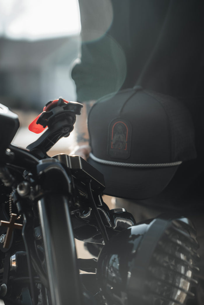 A close-up shows a motorcycle handlebar and headlight, with a person wearing the Born United Private Label Saints and Sinners Hat. The subtle saintly design on the hat catches the sunlight, creating an ethereal lens flare effect in the background.