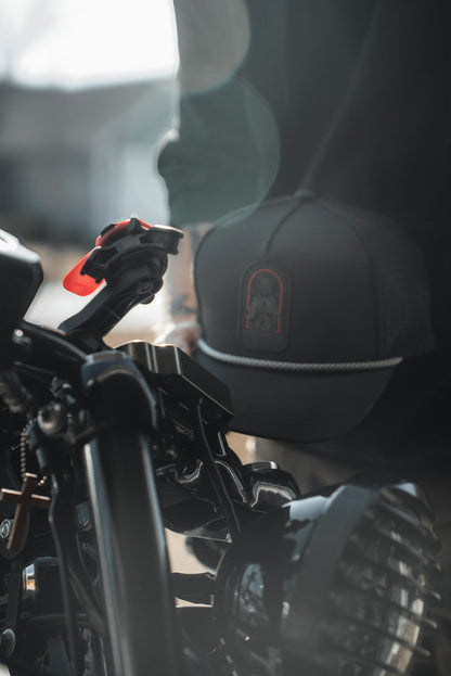 A close-up shows a motorcycle handlebar and headlight, with a person wearing the Born United Private Label Saints and Sinners Hat. The subtle saintly design on the hat catches the sunlight, creating an ethereal lens flare effect in the background.