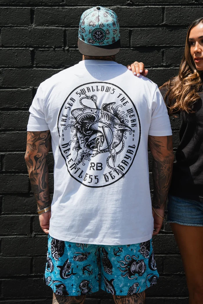 A man in a Relentless Betrayal Sea Swallows The Weak White T-Shirt, blue patterned shorts, and a matching hat stands against a black brick wall as a woman stands beside him with her hand on his back.