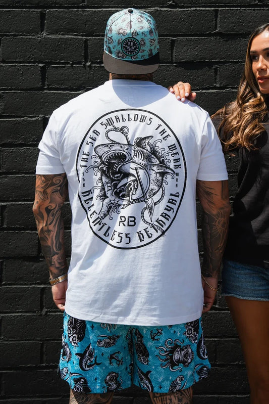 A man in a Relentless Betrayal Sea Swallows The Weak White T-Shirt, blue patterned shorts, and a matching hat stands against a black brick wall as a woman stands beside him with her hand on his back.
