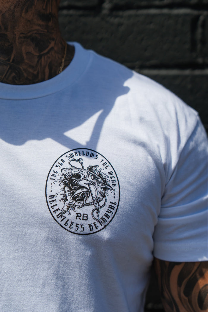 A tattooed individual wears the Sea Swallows The Weak White T-Shirt by Relentless Betrayal—a unisex soft jersey tee with a black and white circular winged figure design and the words “He who swallows the chaos, relentless demon.”.