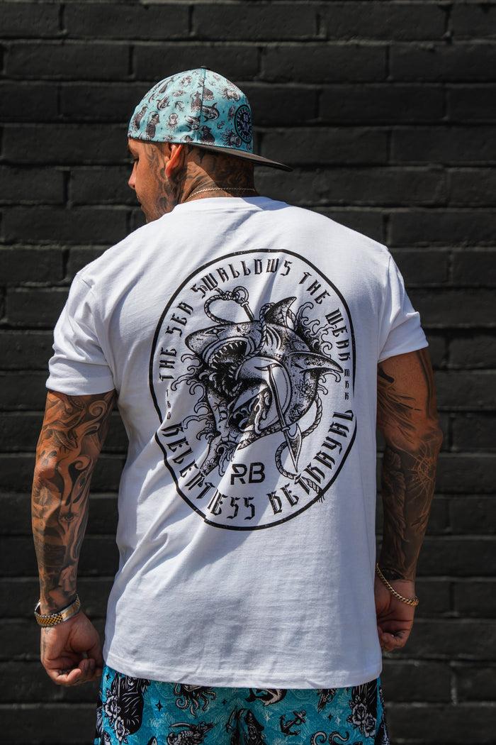 A tattooed man faces away from the camera, wearing the Relentless Betrayal Sea Swallows The Weak White T-Shirt, a bold graphic tee in soft jersey cotton, paired with a blue patterned cap and matching shorts against a black brick wall.