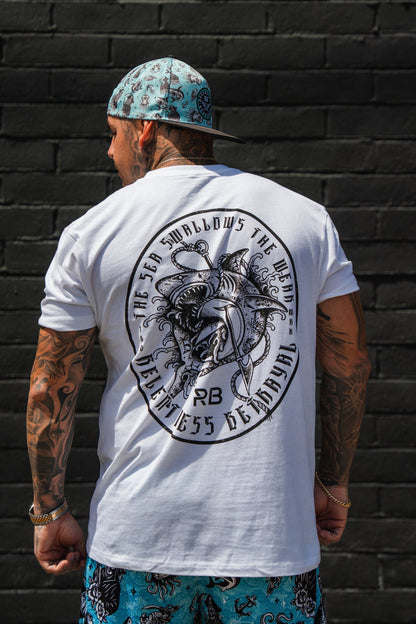 A tattooed man faces away from the camera, wearing the Relentless Betrayal Sea Swallows The Weak White T-Shirt, a bold graphic tee in soft jersey cotton, paired with a blue patterned cap and matching shorts against a black brick wall.