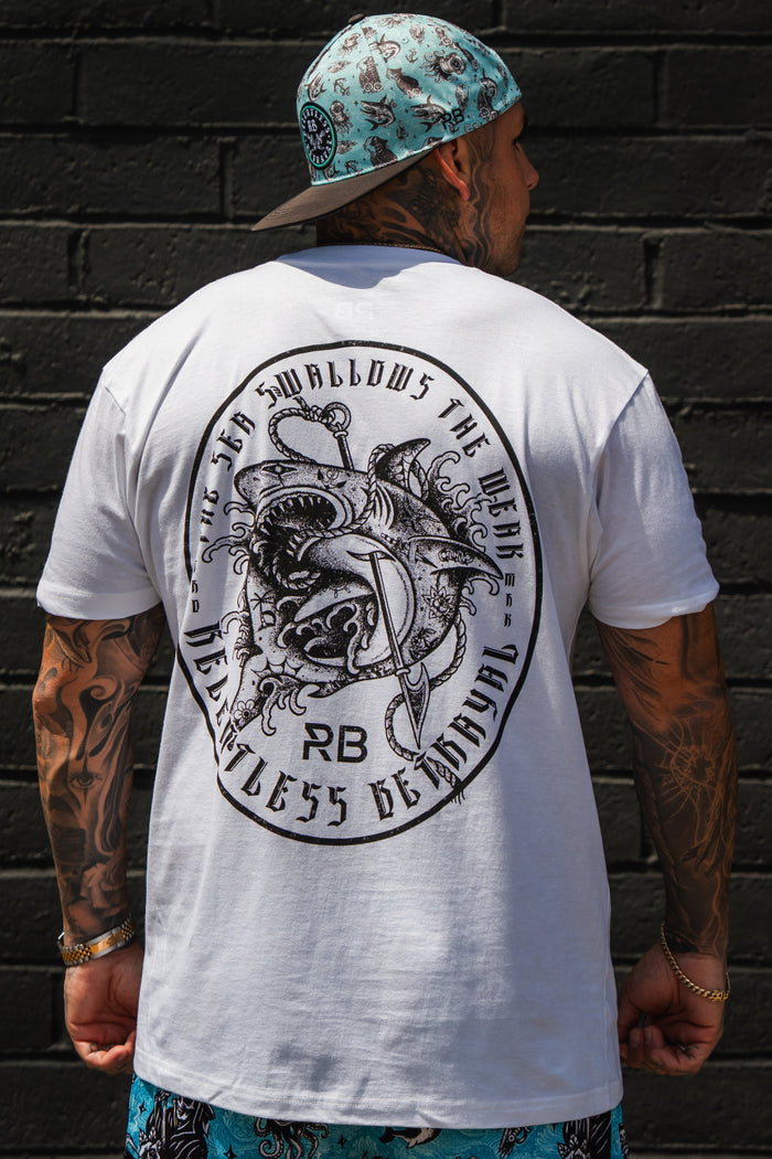 A tattooed person in the Sea Swallows The Weak White T-Shirt by Relentless Betrayal, blue patterned shorts, and a matching blue cap stands before a black brick wall.