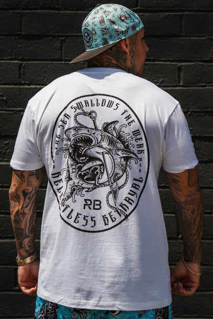 A tattooed person in the Sea Swallows The Weak White T-Shirt by Relentless Betrayal, blue patterned shorts, and a matching blue cap stands before a black brick wall.