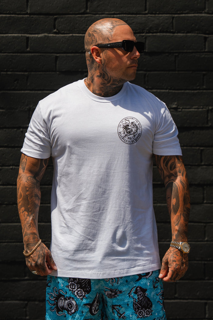 A tattooed man in sunglasses stands against a black brick wall, wearing the Relentless Betrayal Sea Swallows The Weak White T-Shirt with a chest logo and blue patterned shorts.