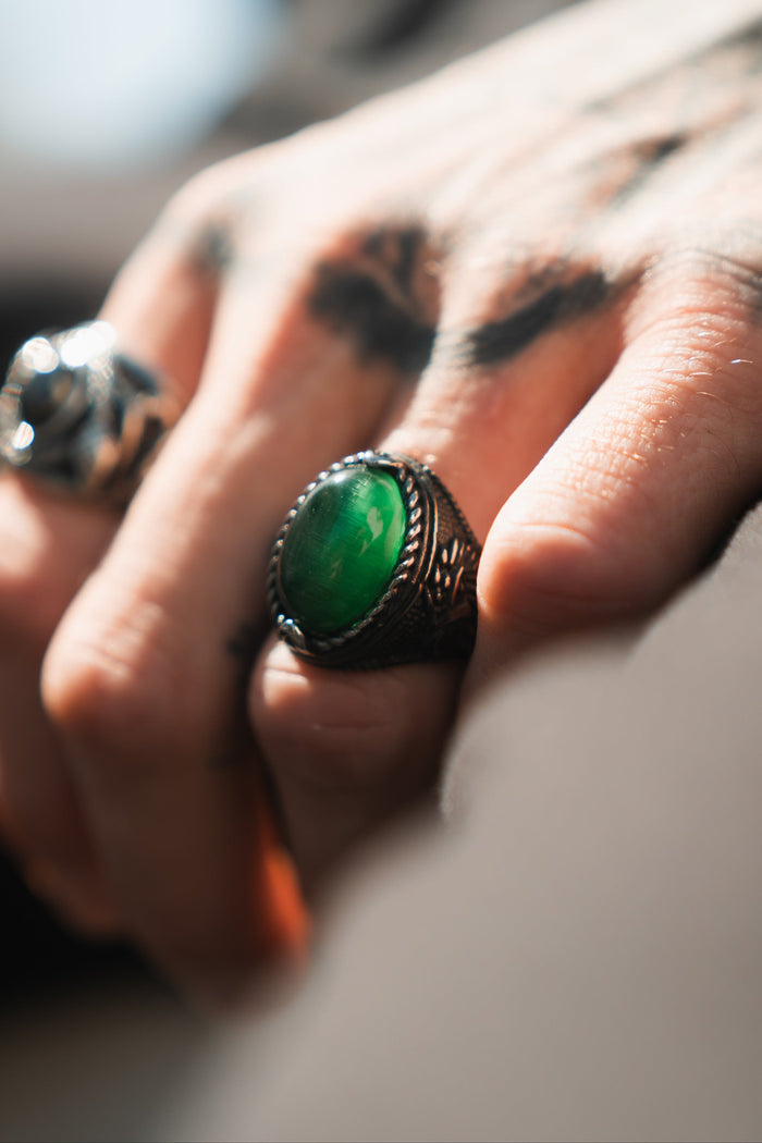 A hand with tattoos rests on a surface, showcasing two rings. Prominently featured is the Relentless Betrayals Serpents Eye Ring with an oval green gemstone, while lighting accentuates the intricate textures and details.