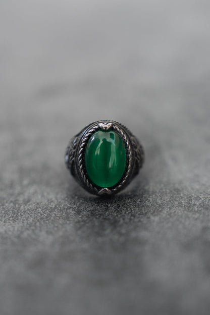 The Serpents Eye Ring from Relentless Betrayal, featuring a large oval green gemstone at its heart and an ornate silver rope-patterned band, rests elegantly on a smooth gray surface.
