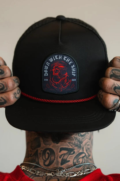 A person holds the brim of a Relentless Betrayal Ship Wreck Snapback, revealing tattoos on their fingers and neck. This black cap features a round patch with a red art design and DOWN WITH THE SHIP phrase. They accessorize with silver rings and a necklace.