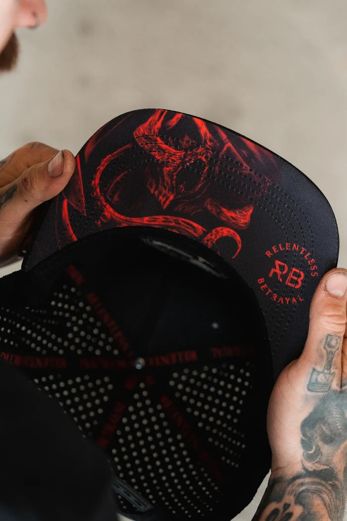 Tattooed hands display the Ship Wreck Snapback - Red from Relentless Betrayal, featuring a black cap with a red demonic illustration under the brim and adorned with RELENTLESS BETRAYAL and RB text for those who love unique style.