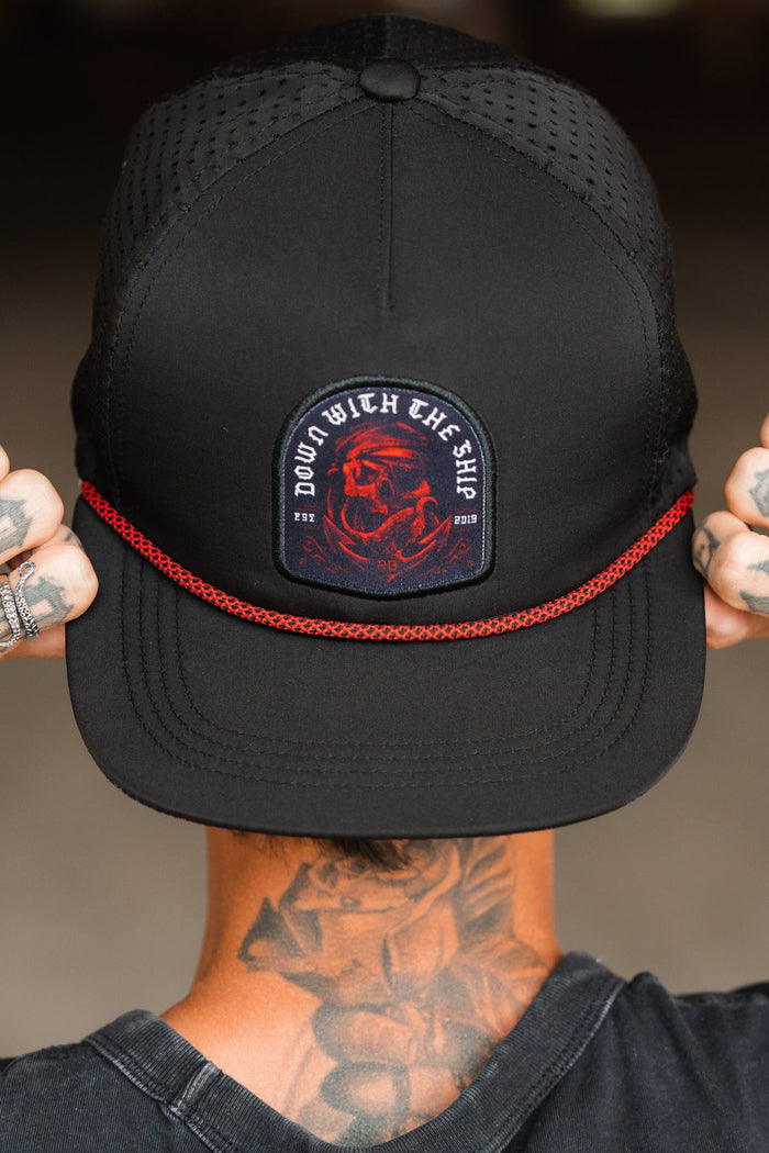The image shows a person from behind wearing the Relentless Betrayal Ship Wreck Snapback - Red, a black cap with a red dragon patch and DOWN WITH THE SHIP in white. The brim features red rope detail, complementing their tattooed neck and hands for a unique style.