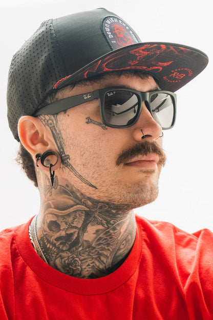 A man wears Relentless Betrayals Ship Wreck Snapback - Red, paired with a red shirt. He sports tattoos on his neck, a mustache, sunglasses, a nose ring, ear gauges, and a black cap with its striking design. His expression is neutral.