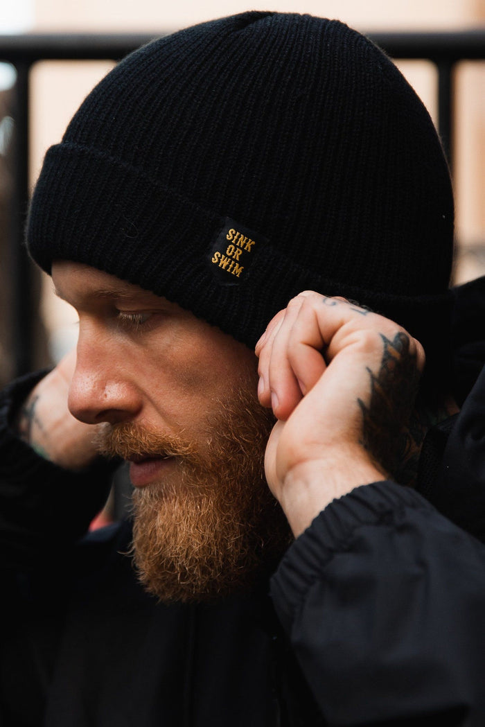 A person with a beard adjusts their Sink or Swim Beanie by Relentless Betrayal, made from 100% acrylic. Theyre in a black jacket, tattoos visible, as the softly blurred background adds an artistic touch.