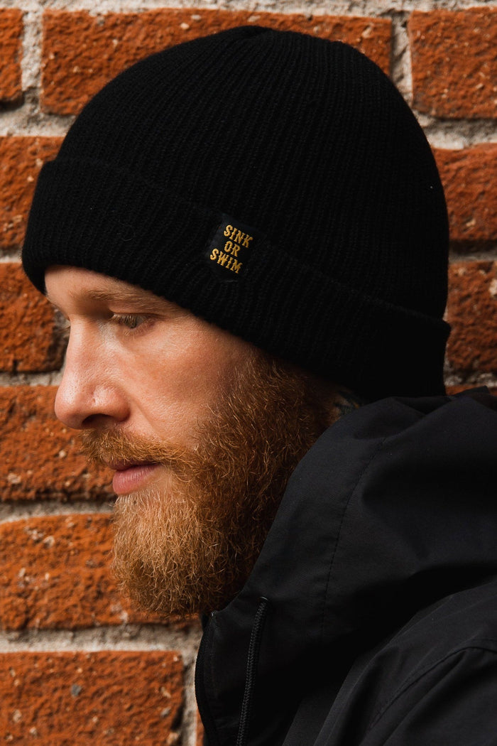 A bearded person wearing the unisex Sink or Swim Beanie by Relentless Betrayal, made of 100% acrylic, and a black jacket is seen in profile. The beanie features a Sink or Swim patch, set against a textured red brick wall.