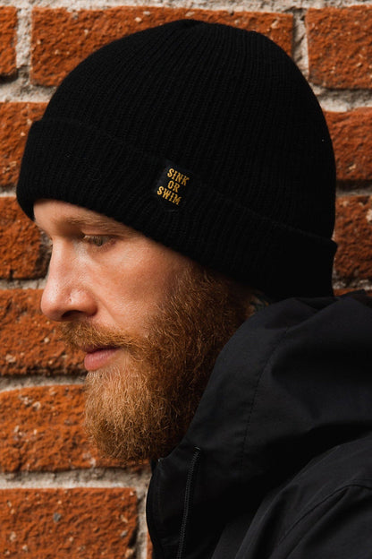 A bearded person wearing the unisex Sink or Swim Beanie by Relentless Betrayal, made of 100% acrylic, and a black jacket is seen in profile. The beanie features a Sink or Swim patch, set against a textured red brick wall.