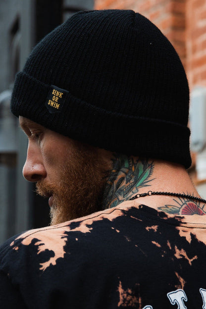 A bearded person showcases a black Sink or Swim Beanie by Relentless Betrayal, made of 100% acrylic, revealing a striking neck tattoo. They wear a black tie-dye shirt and a beaded necklace, standing against a brick wall with a blurred structure in the background.