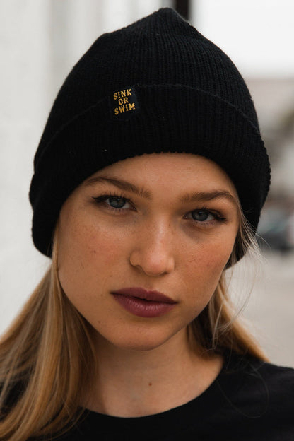 Wearing the unisex Sink or Swim Beanie by Relentless Betrayal, made from 100% acrylic with SINK OR SWIM embroidery, a person with long hair stands against a blurred outdoor backdrop, their expression neutral.