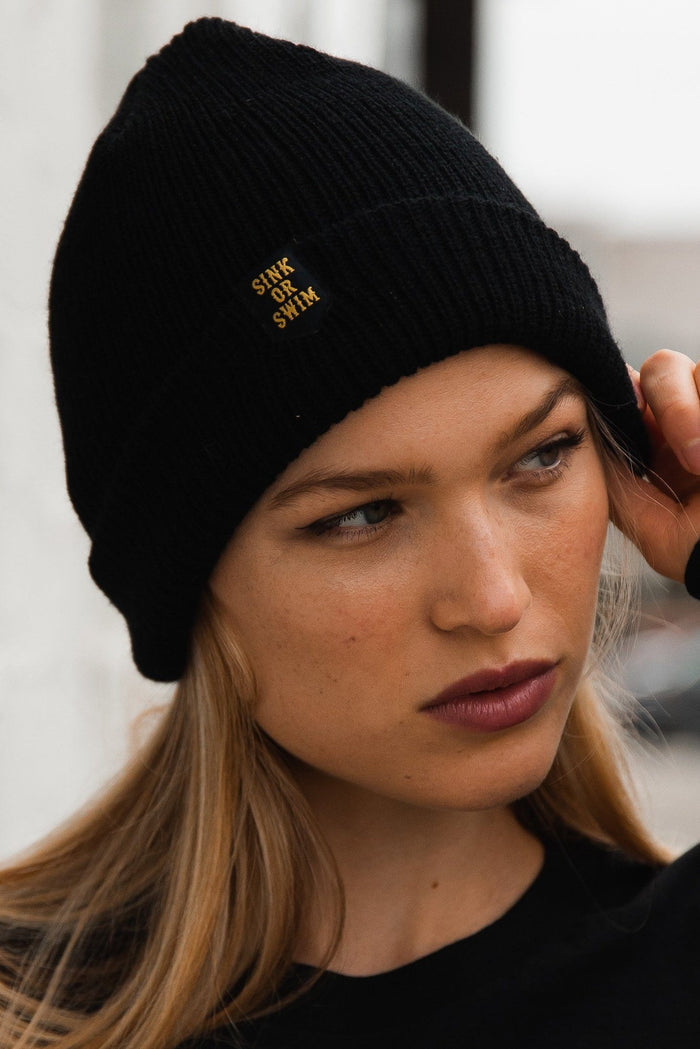 A person in a black shirt, wearing Relentless Betrayal’s unisex Sink or Swim Beanie crafted from 100% acrylic, gazes thoughtfully to the side with flowing long hair against a blurred, mysterious background.