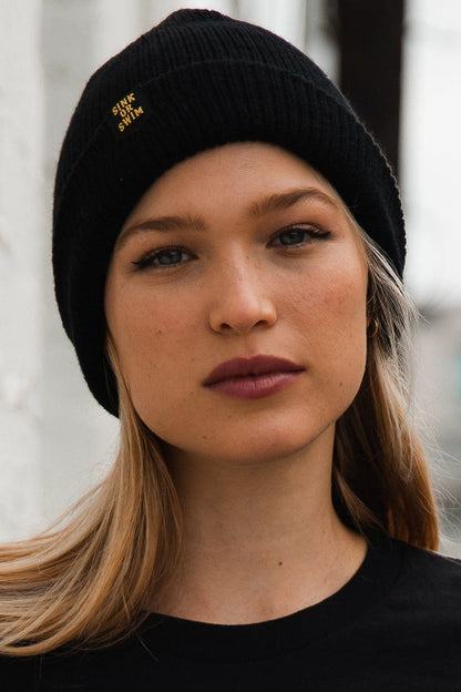 A woman with long hair wears Relentless Betrayals Sink or Swim Beanie, made of 100% acrylic. This unisex beanie features bold white SINK OR SWIM lettering. She gazes straight at the camera with a neutral expression, against a softly blurred background adding depth.