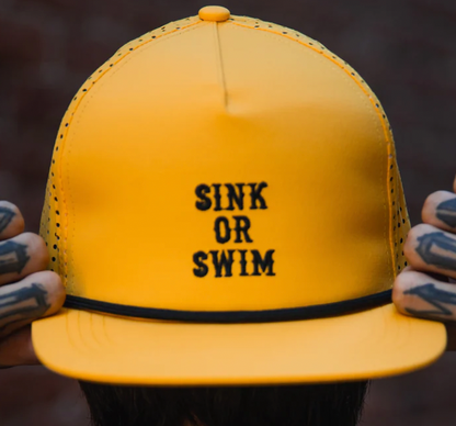 Tattooed hands grip the sides of the Relentless Betrayal Sink or Swim GOLD Snapback, a bold yellow hat featuring "SINK OR SWIM" in black letters on the front.