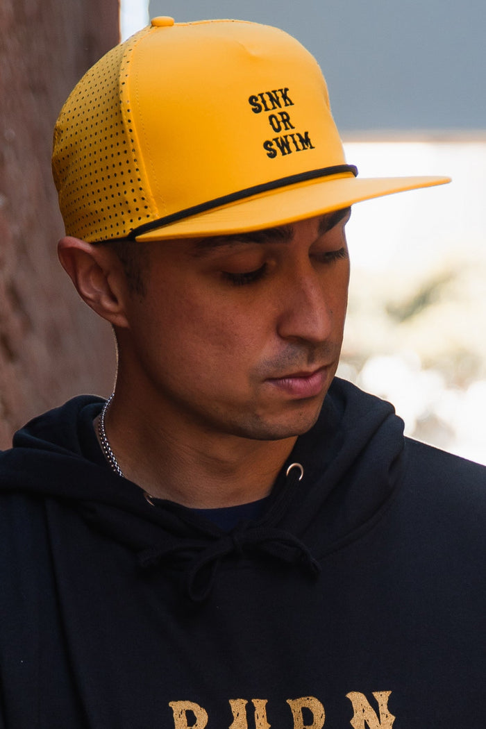 A person in a Relentless Betrayal Sink or Swim GOLD Snapback and a black hoodie looks down near a wall, lit by natural light.