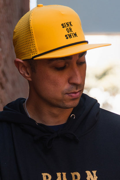 A person in a Relentless Betrayal Sink or Swim GOLD Snapback and a black hoodie looks down near a wall, lit by natural light.