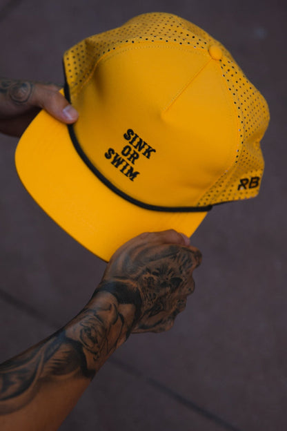A tattooed hand holds the Relentless Betrayal Sink or Swim GOLD Snapback, a bright yellow, water-resistant hat with “SINK OR SWIM” on the front and “RB” on the side, standing out against a muted background.
