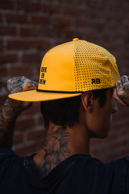A tattooed individual adjusts the Relentless Betrayal Sink or Swim GOLD Snapback, featuring bold black text and RB initials, while standing before a brick wall.