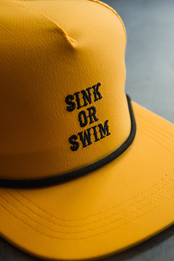 Close-up of the Sink or Swim GOLD Snapback by Relentless Betrayal, featuring black embroidered "SINK OR SWIM" text on the yellow front.