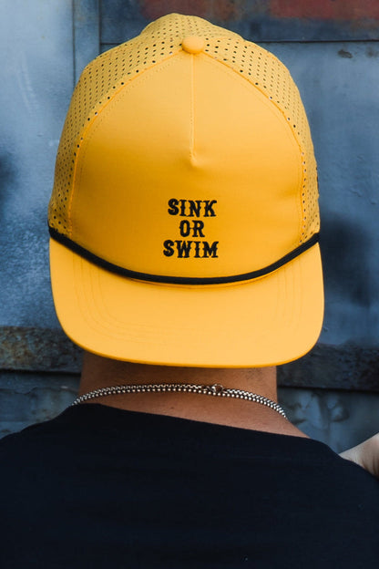 A person, seen from behind, wears the Relentless Betrayal Sink or Swim GOLD Snapback—a bright yellow, water-resistant hat with "SINK OR SWIM" in black, perforated sides, and a silver chain necklace.