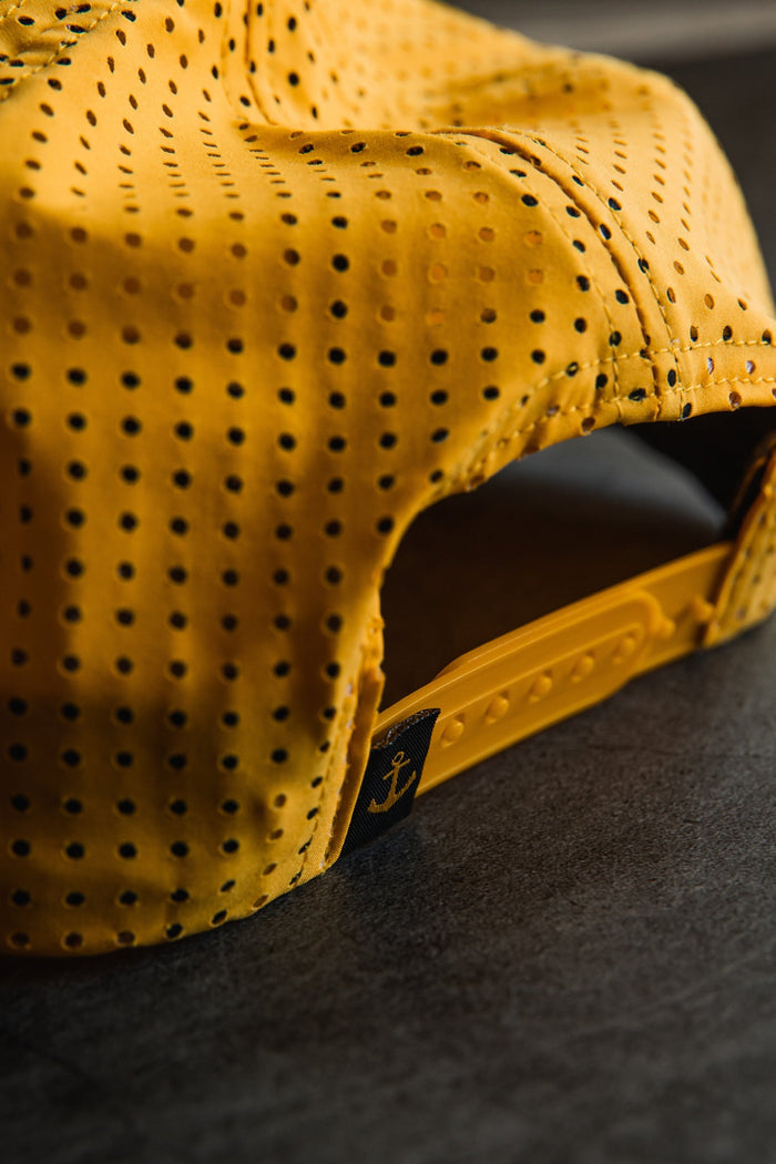 Close-up of the back of the Relentless Betrayal Sink or Swim GOLD Snapback, showing its yellow color, plastic adjustable strap, and a small black tag with a yellow anchor symbol, resting on a dark surface.
