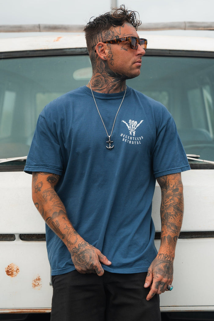 A tattooed man in a Relentless Betrayal Sink or Swim Premium Slate T-Shirt and dark pants stands before a rust-spotted, weathered white vehicle. His sunglasses, necklace, and confidently styled hair exude resilience. The vehicles rust symbolizes perseverance through time.