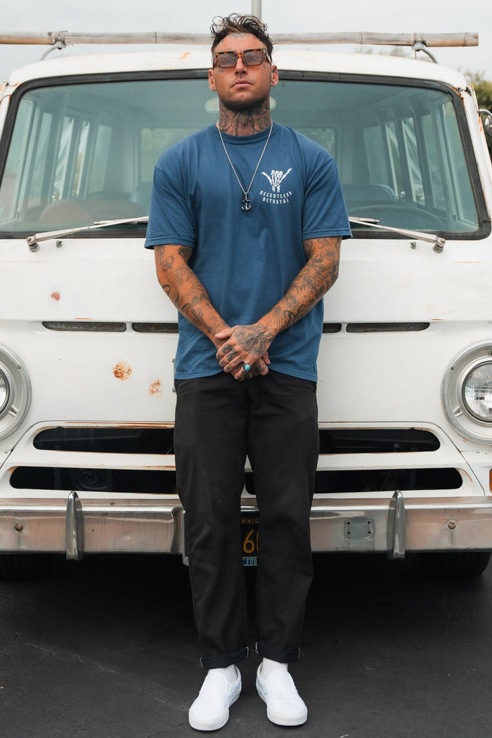 A tattooed man radiates resilience, confidently posing before a vintage white van. He wears sunglasses, black pants, white sneakers, and a Relentless Betrayals Sink or Swim Premium Slate T-Shirt. With a necklace adorning his neck and hands clasped in front, his stance echoes inner strength.