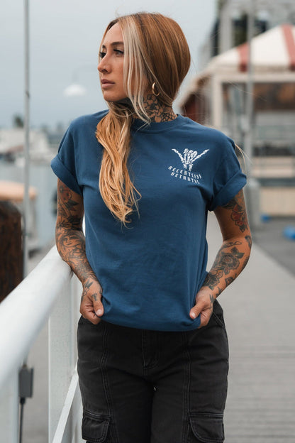 A tattooed woman with long hair, wearing a blue Sink or Swim Premium Slate T-Shirt by Relentless Betrayal, stands on a pier with hands in pockets. She gazes sideways resiliently against an overcast sky and distant buildings.