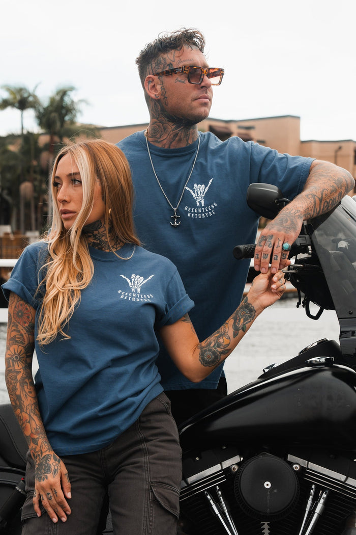 Two tattooed individuals stand by a motorcycle in blue Sink or Swim T-shirts by Relentless Betrayal, featuring white designs with text. The man wears sunglasses and leans on the bike, while the woman rests her hand on his arm. Palm trees and buildings provide the backdrop.