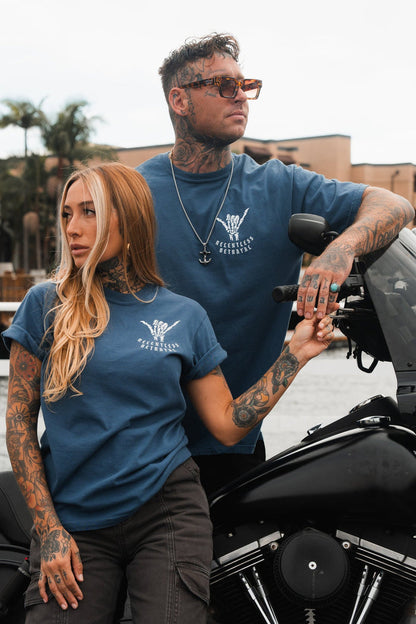 Two tattooed individuals stand by a motorcycle in blue Sink or Swim T-shirts by Relentless Betrayal, featuring white designs with text. The man wears sunglasses and leans on the bike, while the woman rests her hand on his arm. Palm trees and buildings provide the backdrop.