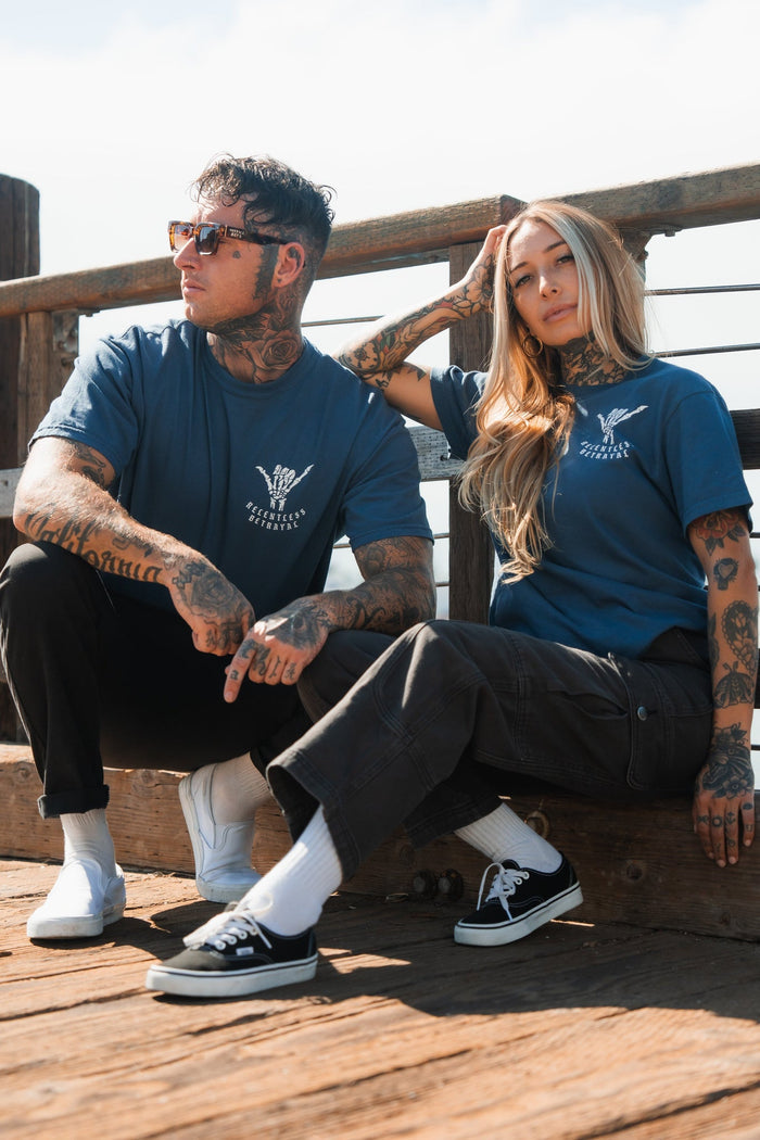 On a wooden deck, two tattooed individuals showcase resilience wearing Sink or Swim Premium Slate T-Shirts from Relentless Betrayal and sunglasses. One confidently sits on a railing while the other squats beside them, with the structure reflecting their inner strength.