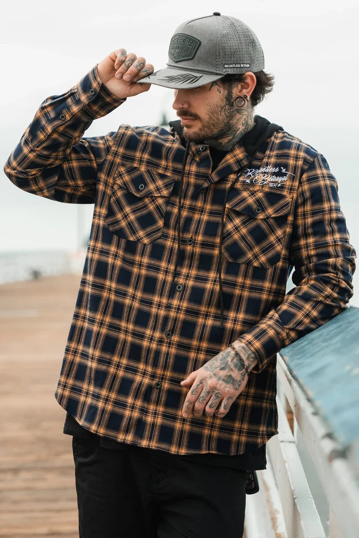 A person in a gray cap and a plaid shirt from Relentless Betrayals Skilled Sailor Premium Hooded Flannel leans on a wooden railing. Outside on a boardwalk, they tilt their cap with one hand, showcasing tattoos on their hand and neck.