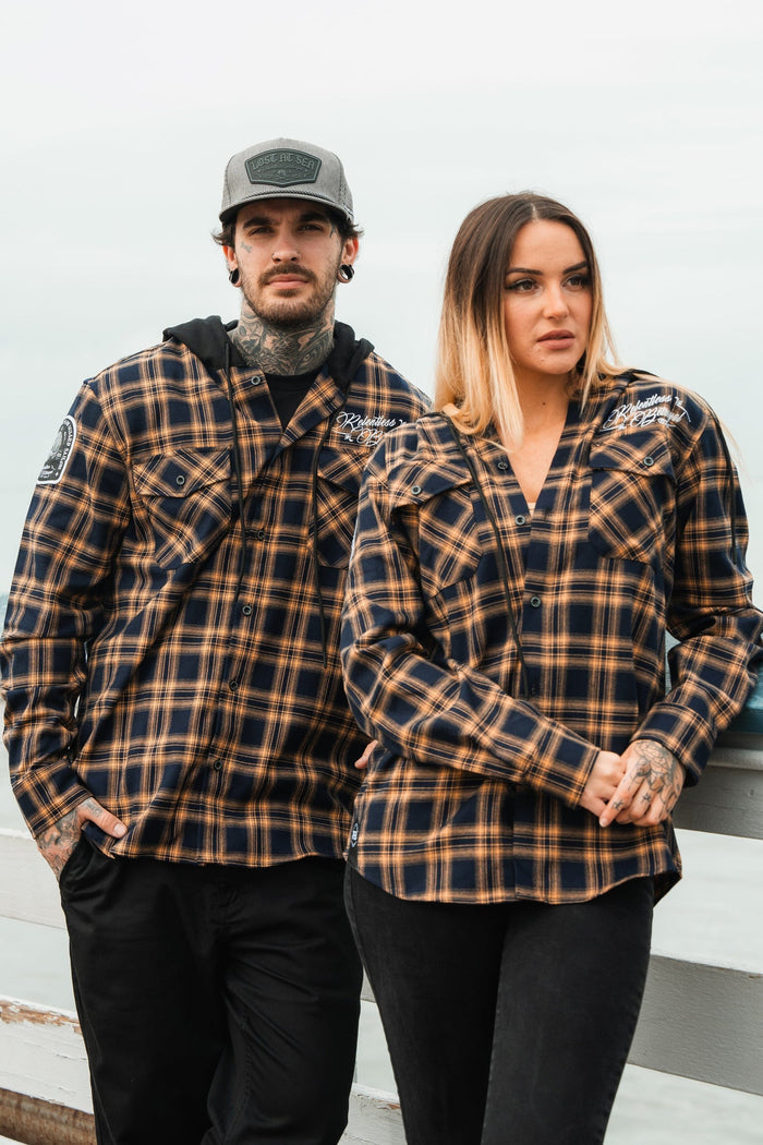 A man and woman in matching Relentless Betrayals Skilled Sailor Premium Hooded Flannels and dark pants stand side by side. The man, wearing a cap, has his arm on a railing as the woman stands with her hands by her sides under a pale sky outdoors.
