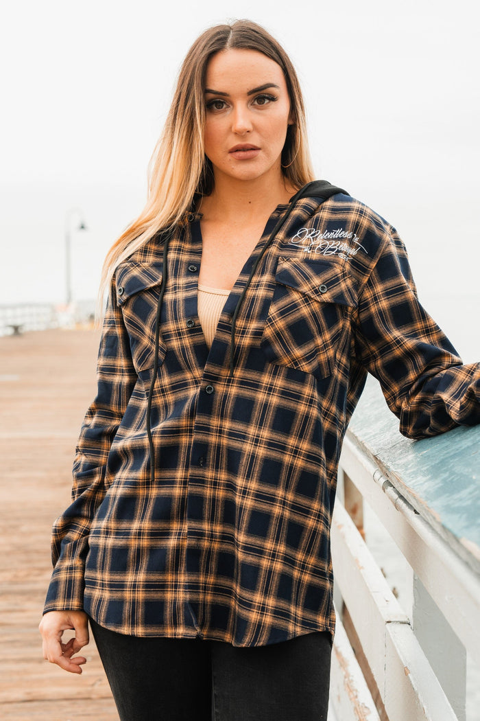 A person with long hair stands on a wooden boardwalk near a railing, wearing the Skilled Sailor Premium Hooded Flannel by Relentless Betrayal—a black and brown plaid shirt over a light-colored top—with a pale, overcast sky in the background.