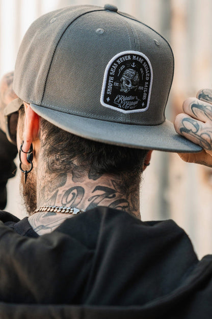 A person with tattoos on their neck adjusts a Relentless Betrayal Skilled Sailor Snapback, gray with a Smooth Seas Never Made Skilled Sailors patch. They wear a black jacket and earrings, facing away from the camera.