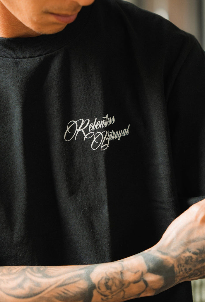 A person wearing a black Skilled Sailor Tee by Relentless Betrayal features Rebellious Betrayal in cursive. Their tattooed arms and partially visible face reflect overcoming challenges, embodying the spirit of a seasoned sailor braving stormy seas.