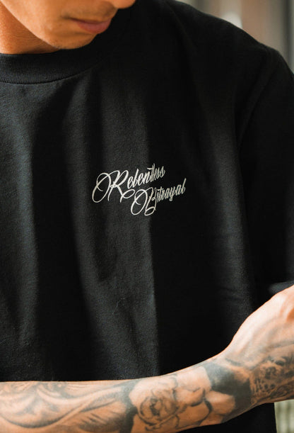 A person wearing a black Skilled Sailor Tee by Relentless Betrayal features Rebellious Betrayal in cursive. Their tattooed arms and partially visible face reflect overcoming challenges, embodying the spirit of a seasoned sailor braving stormy seas.