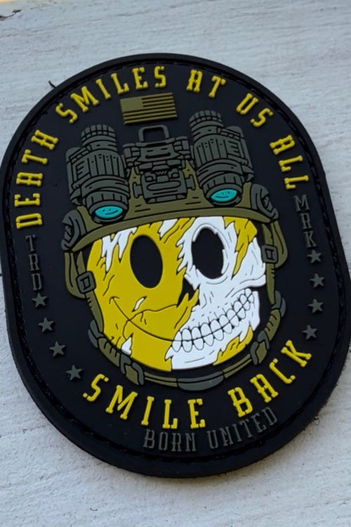 The Smile Back PVC Patch by Born United Private Label shows a helmeted face—half yellow smiley, half skull with night vision goggles—and the words: Death smiles at us all, smile back. Born United—a symbol of stoic resilience.