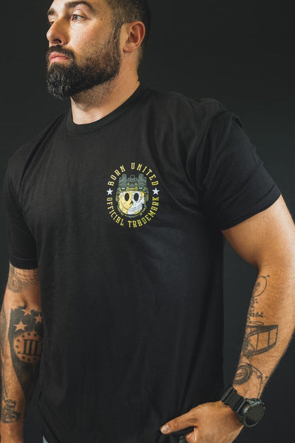 A bearded, tattooed man in a black Born United Private Label "Smile Back" tee and light jeans stands against a black background, gazing confidently to the side with one hand in his pocket.