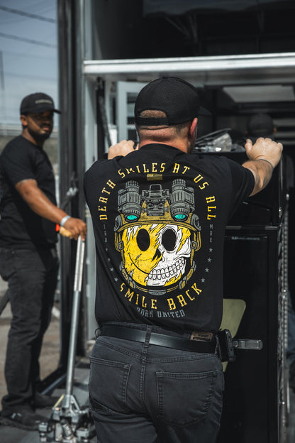 Wearing Born United Private Label’s Smile Back t-shirt featuring a graphic skull and the phrase “DEATH SMILES AT US ALL, SMILE BACK,” one person pushes equipment outdoors as another assists nearby.