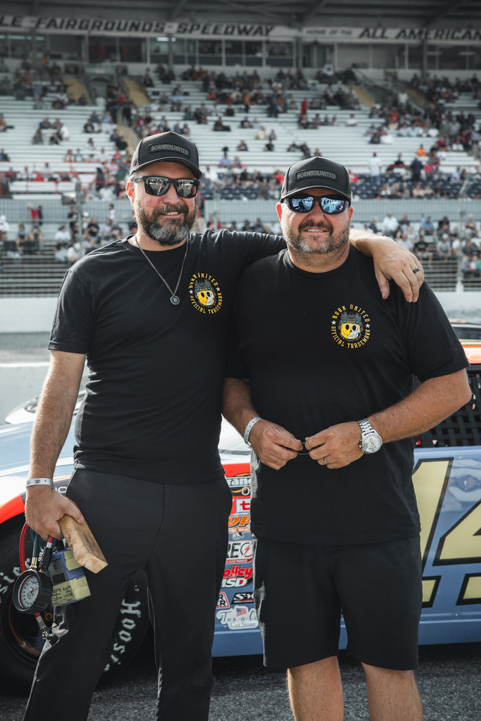 Two men in Born United Private Label’s “Smile Back” apparel—matching tees, caps, and sunglasses—stand smiling together in front of a race car at a speedway, with grandstands full of spectators behind them.